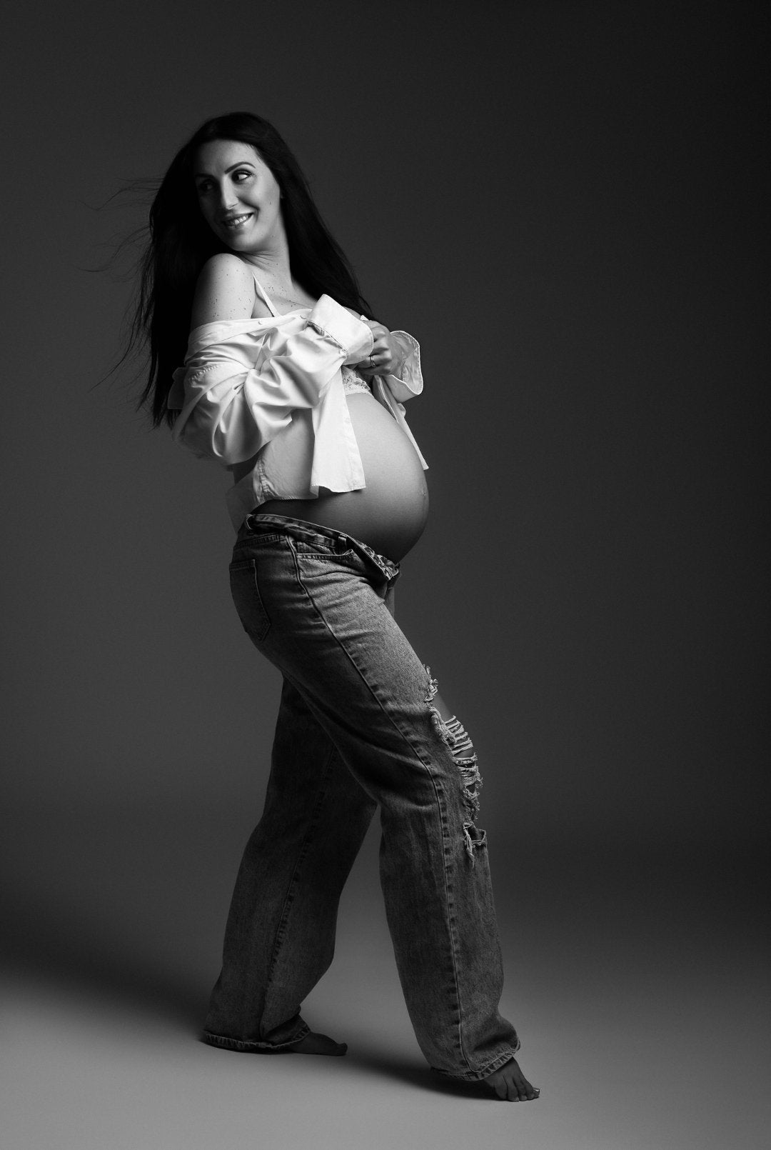 Femme enceinte en jean et chemise blanche ouverte, portrait noir et blanc en studio sur fond gris